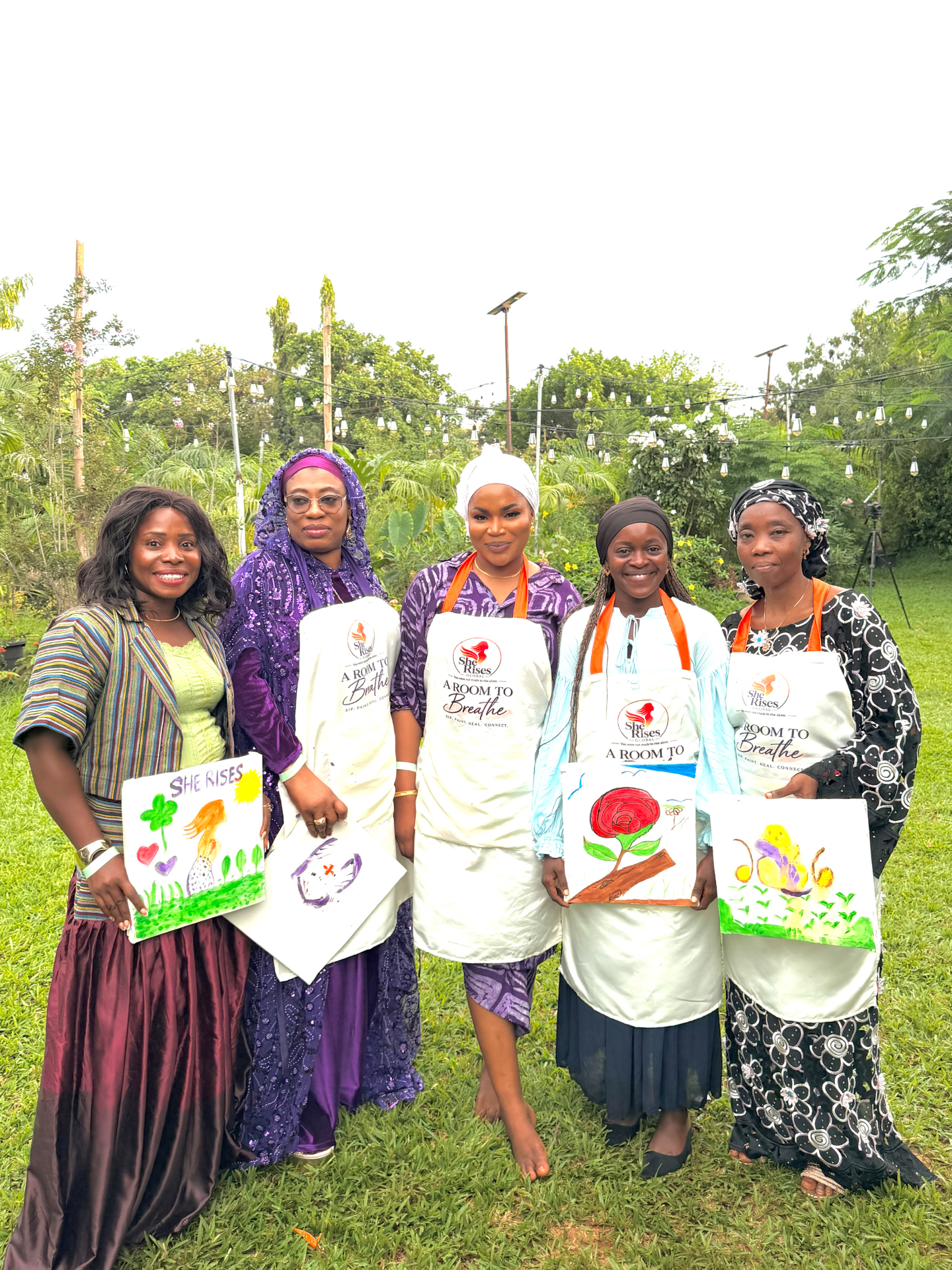 Participants of A Room to Breathe holding their paintings and wearing She Rises aprons